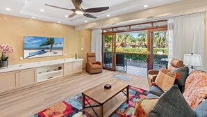 A spacious living room with a ceiling fan, a large window with a view of a garden, a sofa with cushions, and a coffee table. - Stylish 2-Bedroom Beach Condo with Ocean-View Deck, Lush Pool Area & 2-Minute Walk to Flamingo Beach (Cabo Velas)
