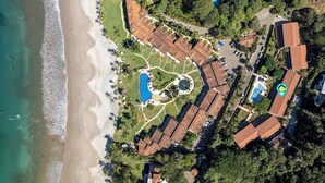An aerial view of a resort with a beach, a swimming pool, and a tennis court. - Stylish 2-Bedroom Beach Condo with Ocean-View Deck, Lush Pool Area & 2-Minute Walk to Flamingo Beach (Cabo Velas)
