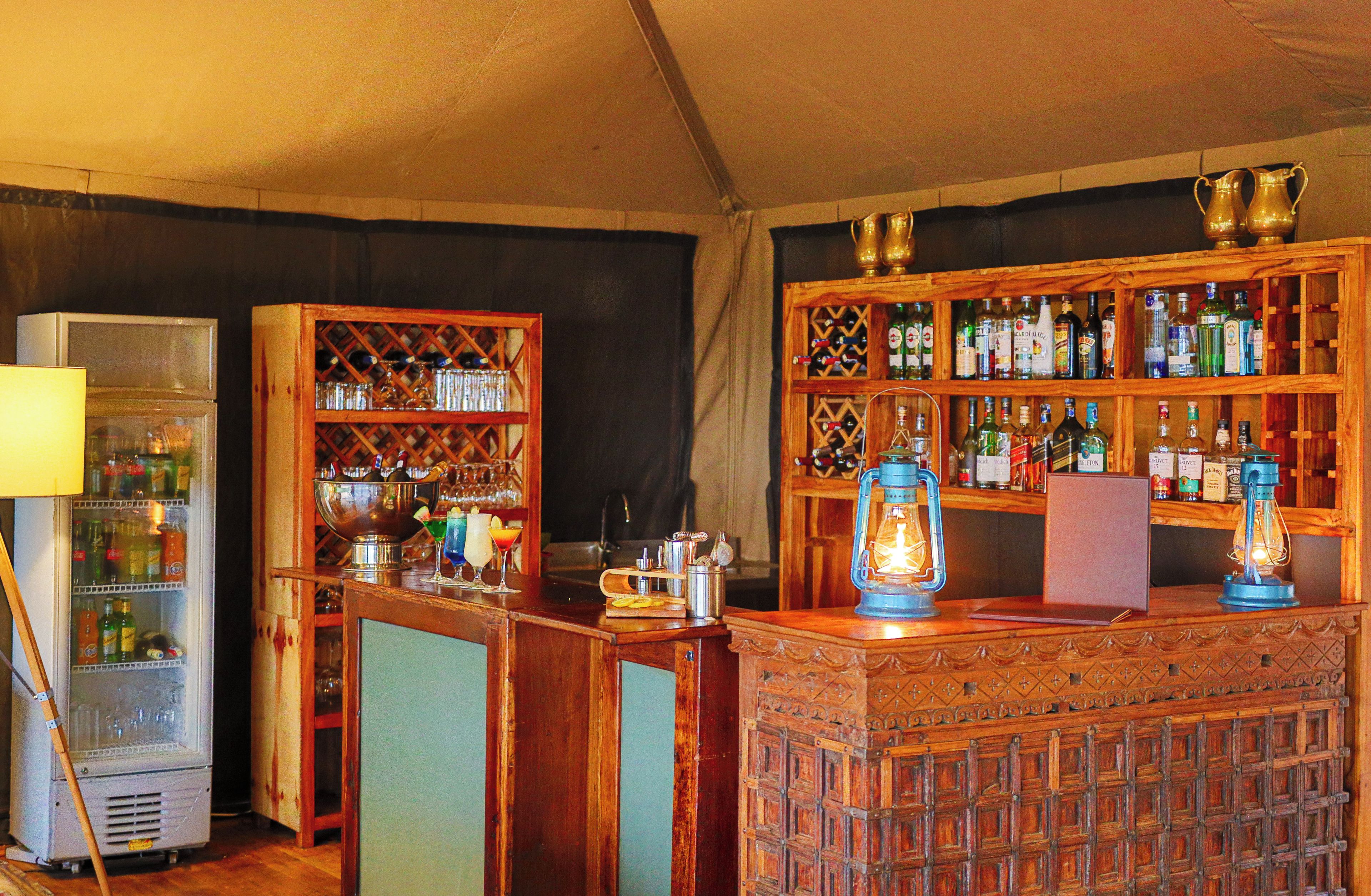 Uma área de bar com design vintage, incluindo um balcão de madeira, prateleiras com garrafas de bebidas e um candeeiro antigo.