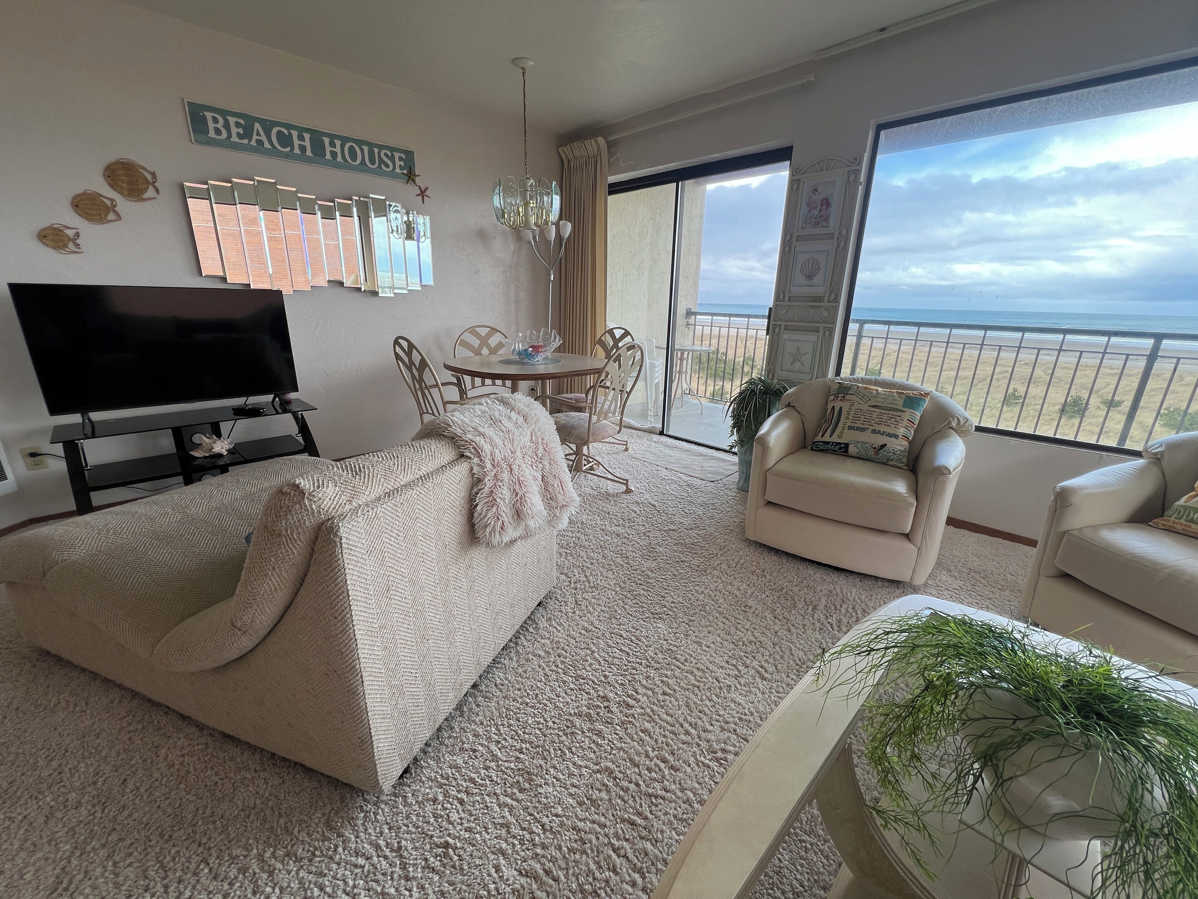 Uma sala de estar com uma televisão de tela plana grande, um sofá, duas poltronas, uma mesa de centro e uma varanda com vista para a praia.