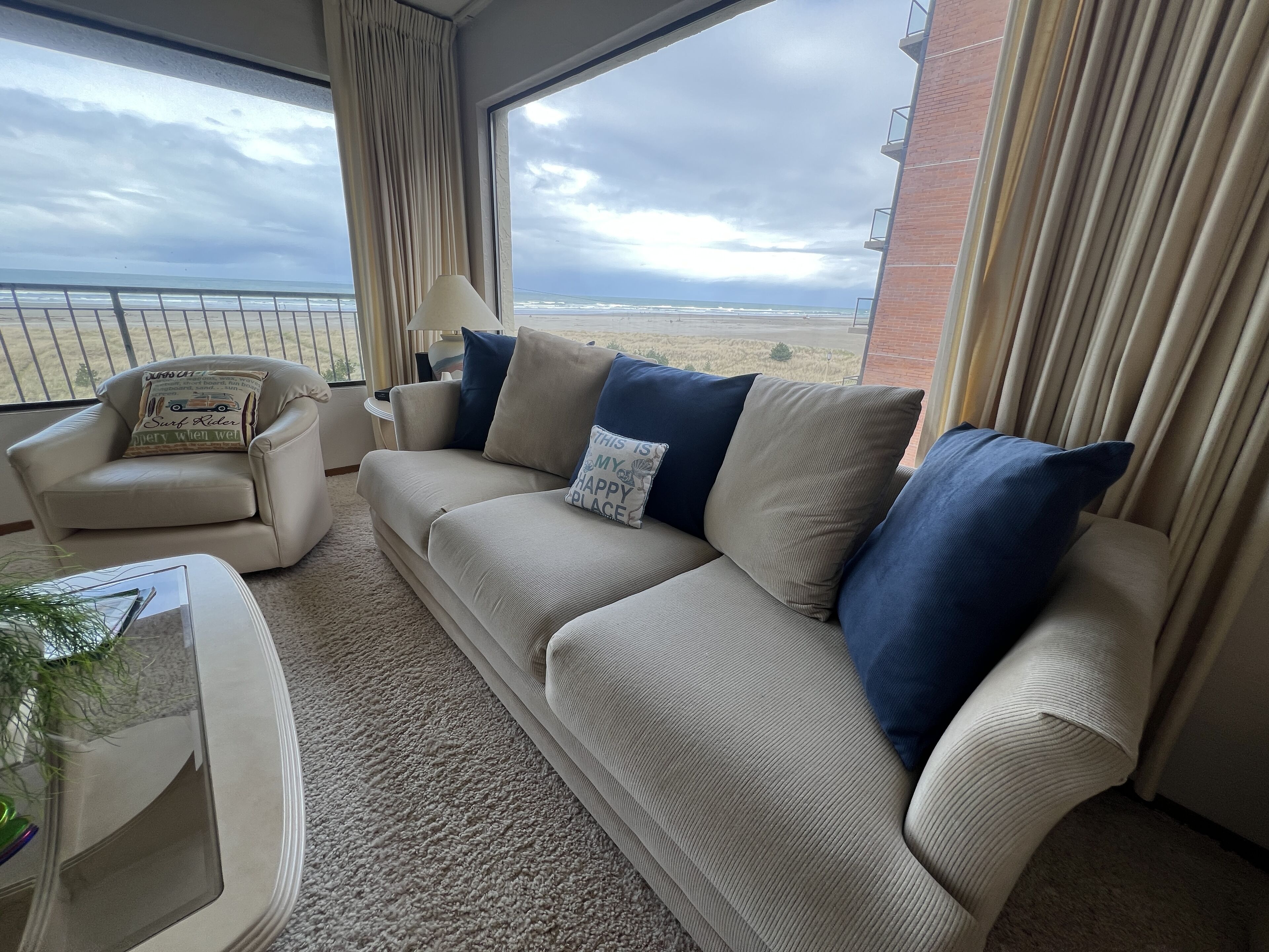 A living room with a beige sofa, a chair, and a small table. There is a view of the beach through large windows.