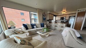 A modern living room with a sofa, armchairs, and a coffee table. There is a kitchen area with white cabinets and a refrigerator. - Beachfront Condo 3rd Floor Corner Unit sleeps 4, Spectacular Panoramic Ocean Vie (Seaside)
