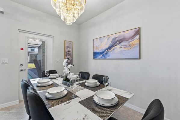 Un área de comedor con una mesa rectangular dispuesta para seis personas, rodeada de sillas negras, una araña y un cuadro abstracto de gran tamaño en la pared.