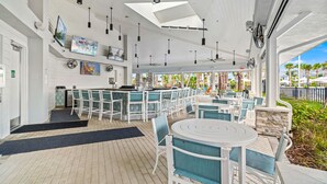Ein überdachter Essbereich im Freien mit weißen Tischen und Stühlen, eine Bar und Deckenventilatoren.