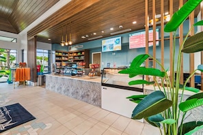 A modern hotel lobby with a reception desk, a seating area with orange chairs, and a large plant in the foreground.