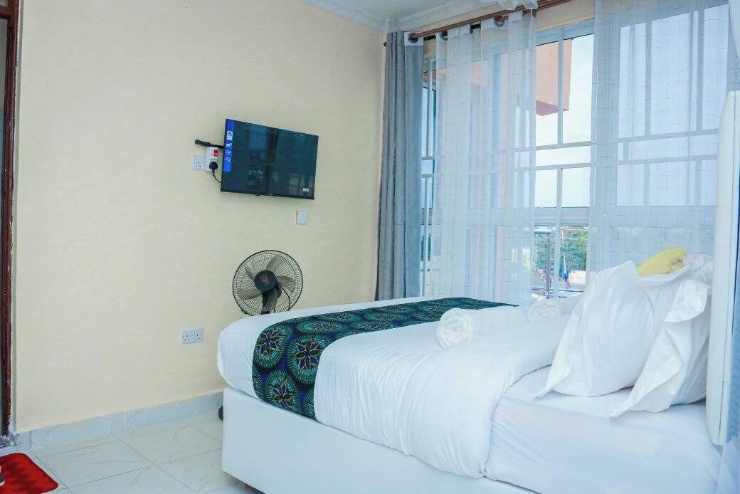 A hotel room with a bed, a TV mounted on the wall, a fan, and a window with curtains.