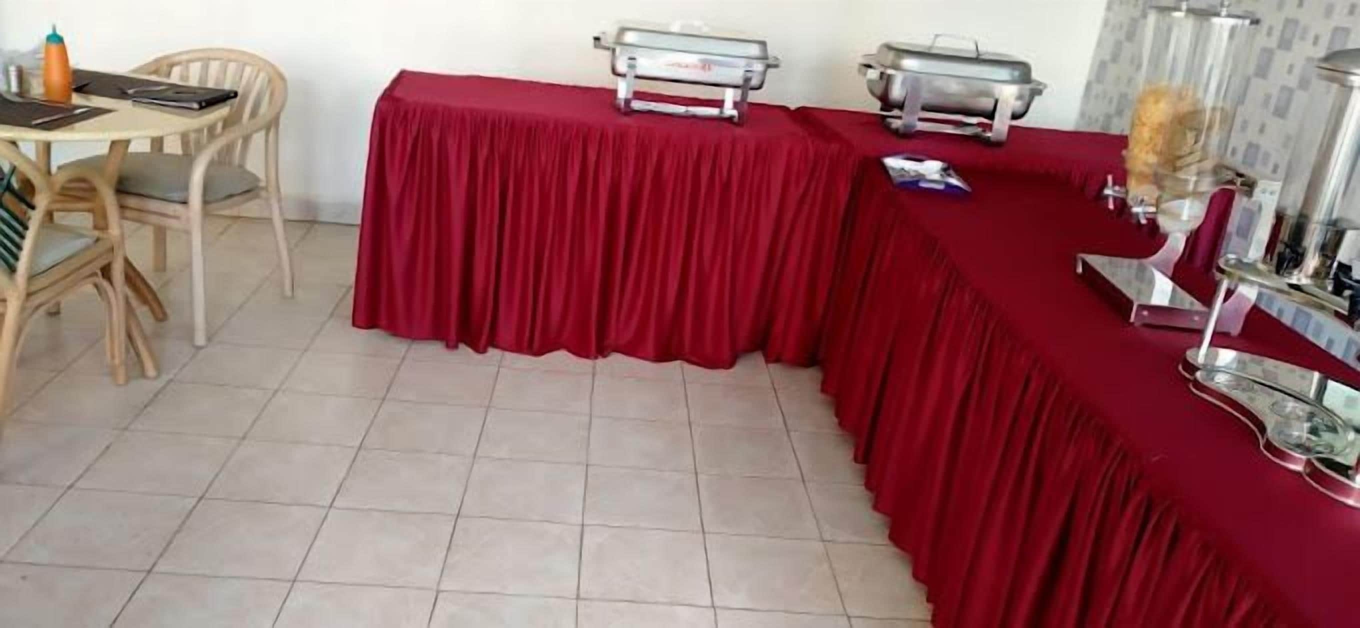 Un comedor con una mesa cubierta con mantel rojo, sillas y equipo para servicio de comida tipo bufé.