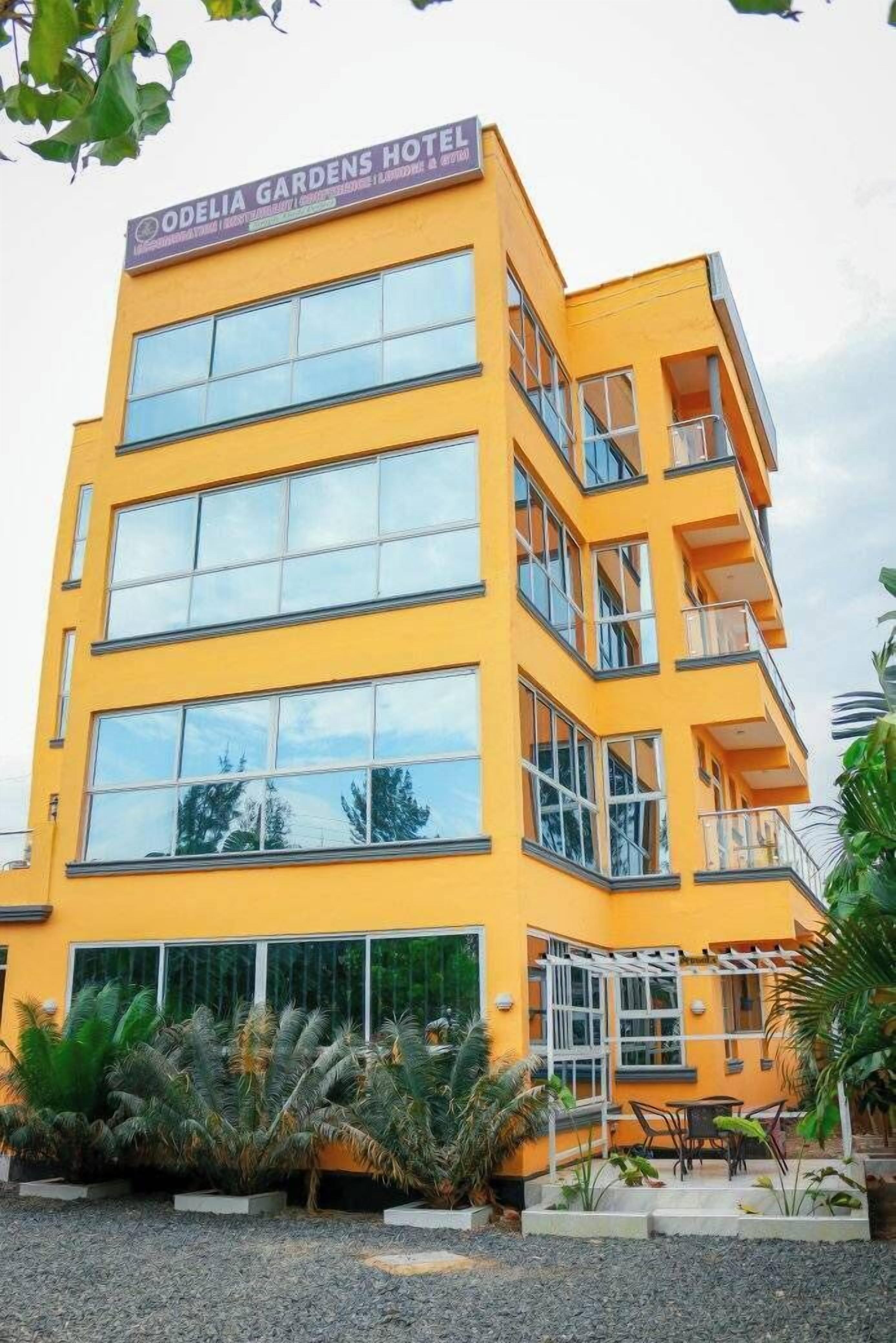 A multi-story hotel with a yellow facade, large windows, and a sign reading "Odelia Gardens Hotel".