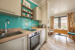 A kitchen with teal cabinetry, a stainless steel sink, a built-in oven, and a wooden countertop. There is a dining area with a table and chairs, and a window with curtains.