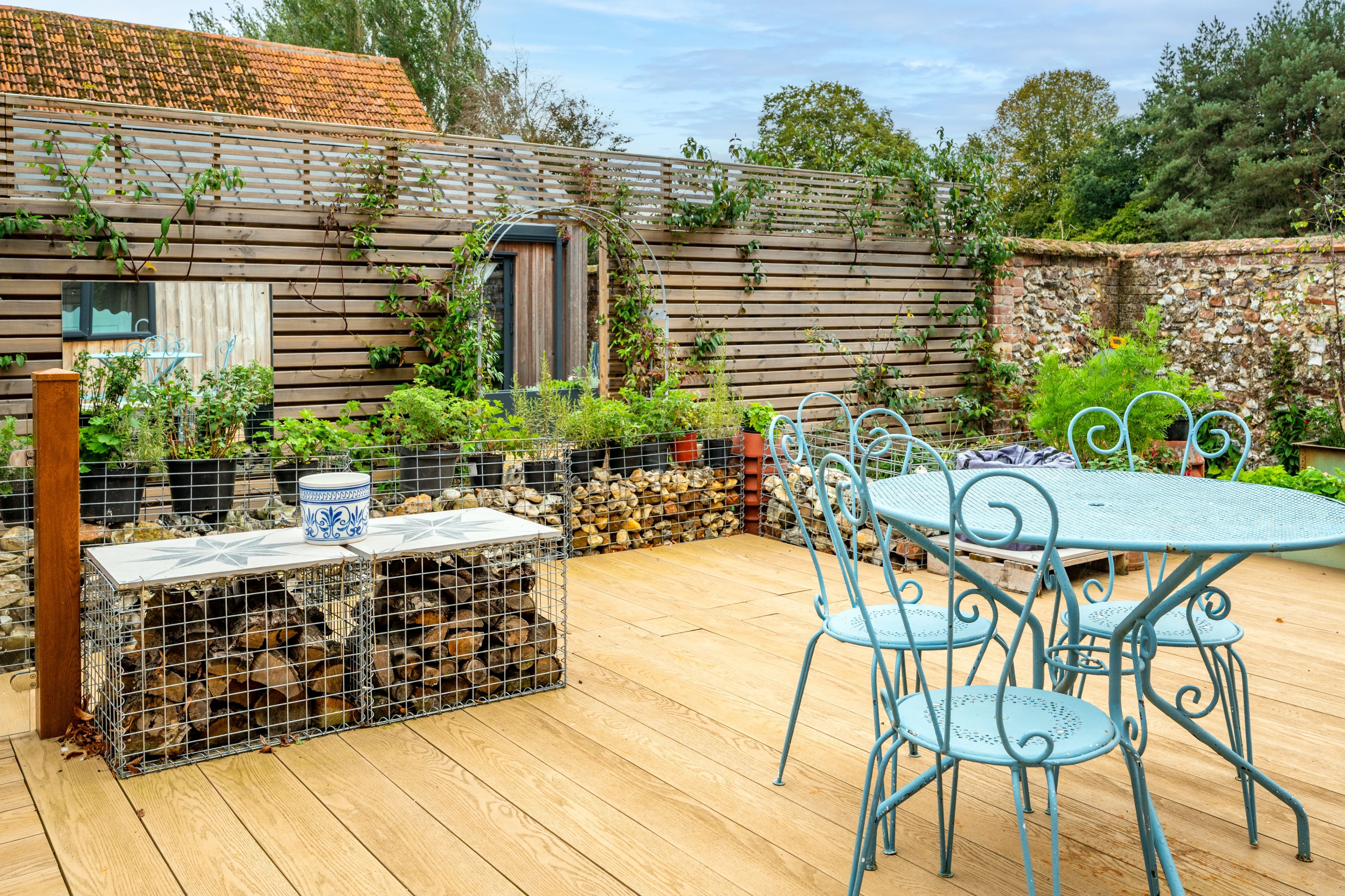 Eine Holzterrasse mit einem Metalltisch und Stühlen, umgeben von Blumentöpfen und einer Steinsäule.