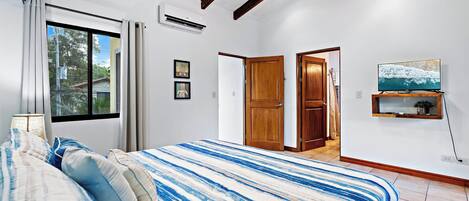 A bedroom with a bed, a window, an air conditioner, and a TV on the wall.