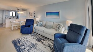 A modern living room with a white sofa, blue armchairs, a patterned rug, and a kitchen in the background. - Seaside 407 | Balcony, Pool, Grill Area + Direct Beach Access (Fort Myers Beach)