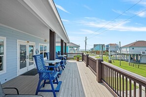 A covered outdoor deck with blue benches and tables, overlooking a grassy area with buildings in the background. - Outlaw's Inn - Gulf View, fenced yard, outdoor bar - 3 Bedroom (Crystal Beach)
