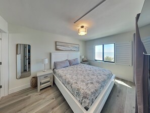 Un dormitorio con cama, mesitas de noche, un espejo, una ventana con contraventanas y una lámpara en el techo.
