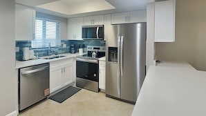 A modern kitchen with white cabinets, stainless steel appliances, a microwave, a refrigerator, and a window with blinds.