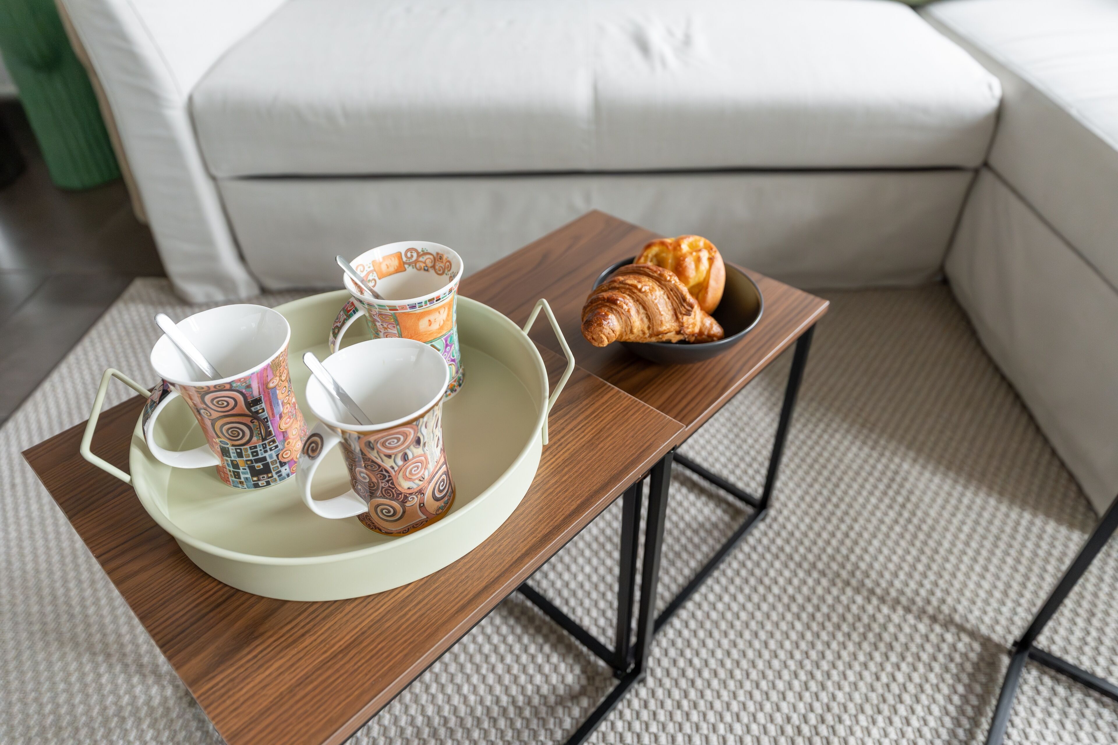 Un plateau de petit-déjeuner avec des tasses et un croissant, posé sur une table en bois dans une pièce où se trouve un canapé blanc.