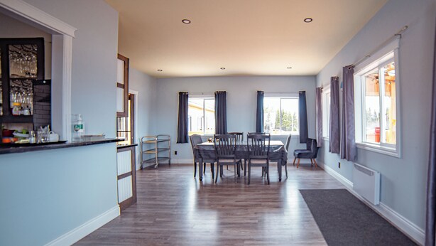 A spacious dining area with a long table and chairs, a kitchen counter with various items, and large windows with curtains. - HOTELLERIE PUAMUN (Aguanish)