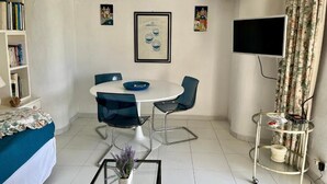 A small, well-lit room with a bed, a round table with chairs, a TV, and a bookshelf.