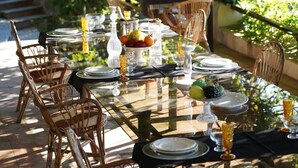 A dining table set outdoors with wicker chairs, clear glassware, and a fruit bowl.