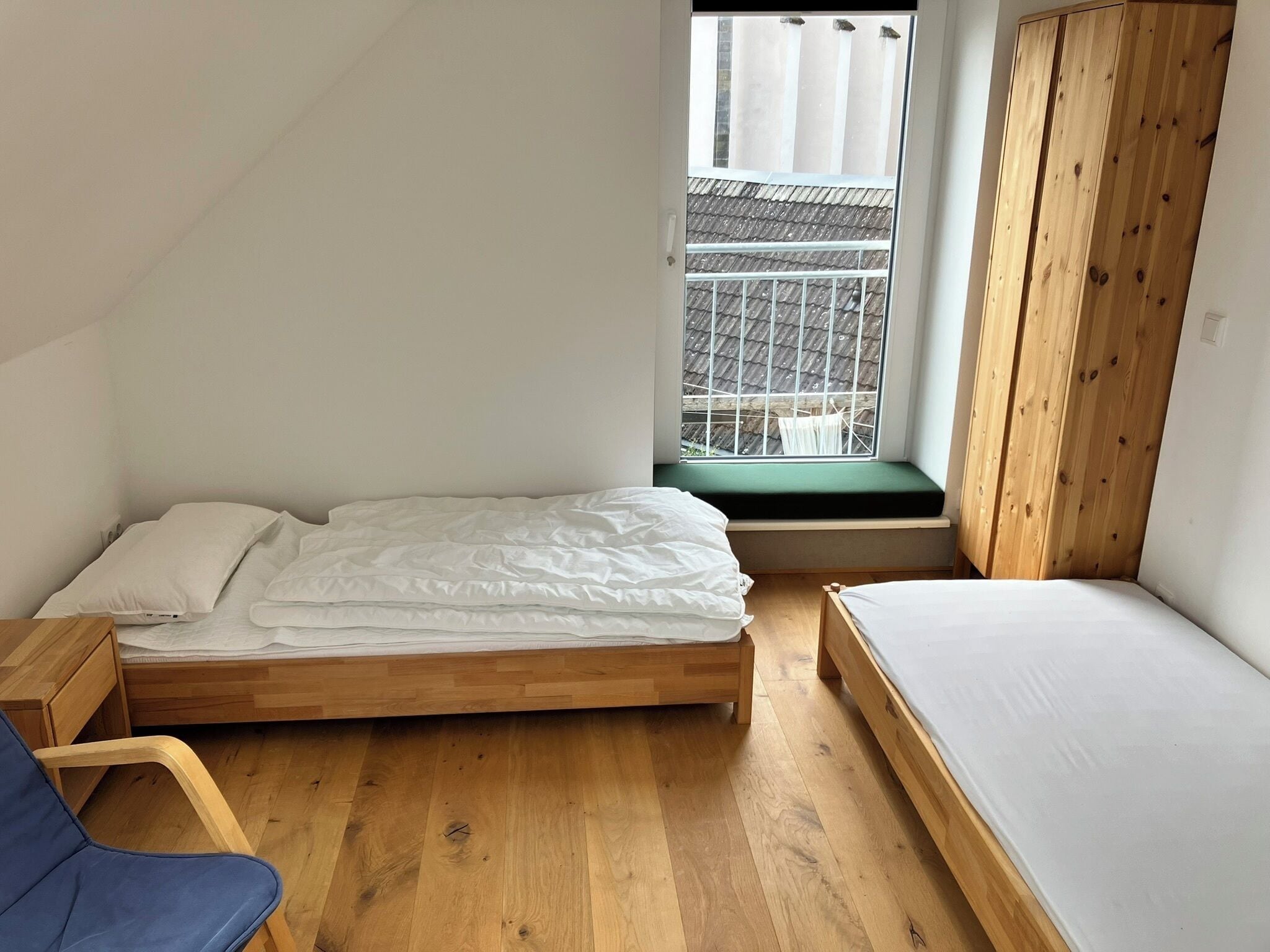 A small, single-room apartment with a wooden floor, a bed with white bedding, a window with a view of a building, and a wooden wardrobe.