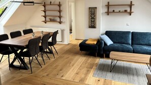 Moderni olohuone, jossa on ruokailutila, sininen sohva ja puuhyllyt.