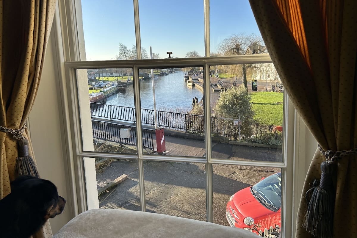A view through a window with curtains, featuring a river, a bridge, and a red car parked outside.