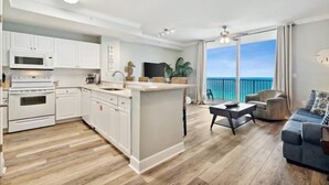 Une cuisine moderne avec des meubles blancs et un îlot central, un coin repas avec une table et des chaises, et un balcon offrant une vue sur l’océan.