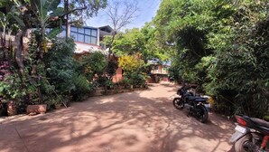 Ein gepflasterter Weg führt zu einem Gebäude, das von üppigem Grün umgeben ist, mit einem abgestellten Motorrad.