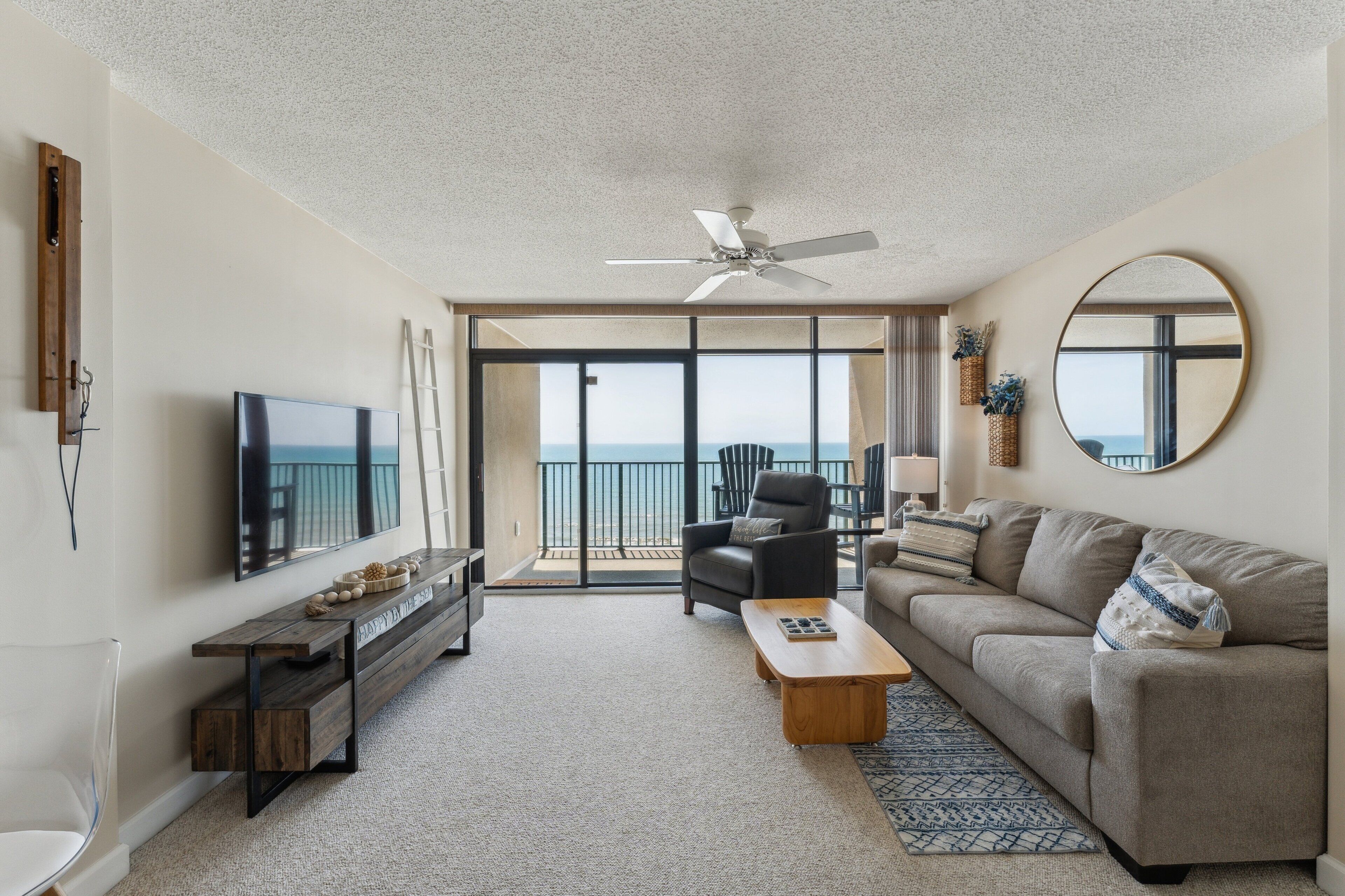 A modern living room with a large TV, a ceiling fan, a sofa, a coffee table, and a view to the outside.