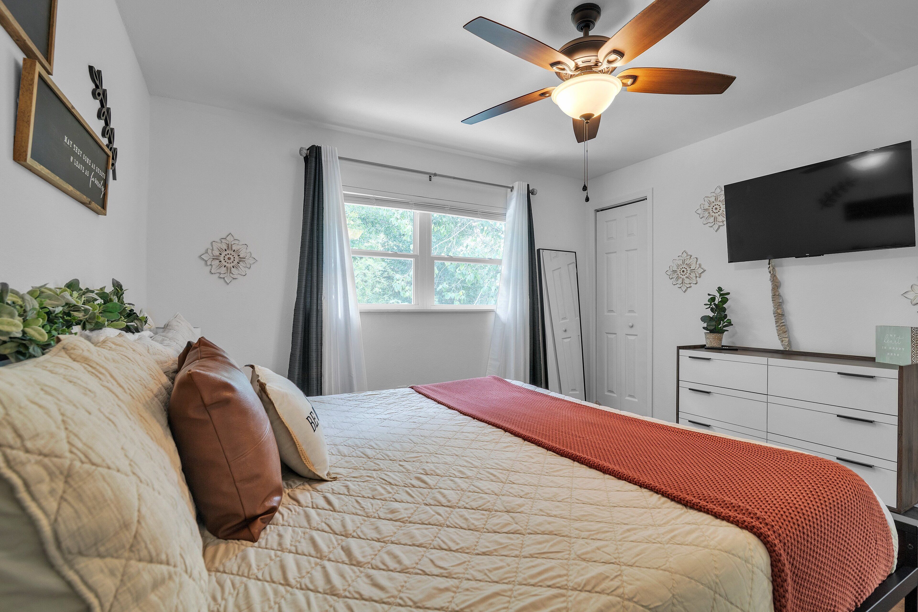 A bedroom with a bed, a ceiling fan, a TV, a window with curtains, and a dresser.