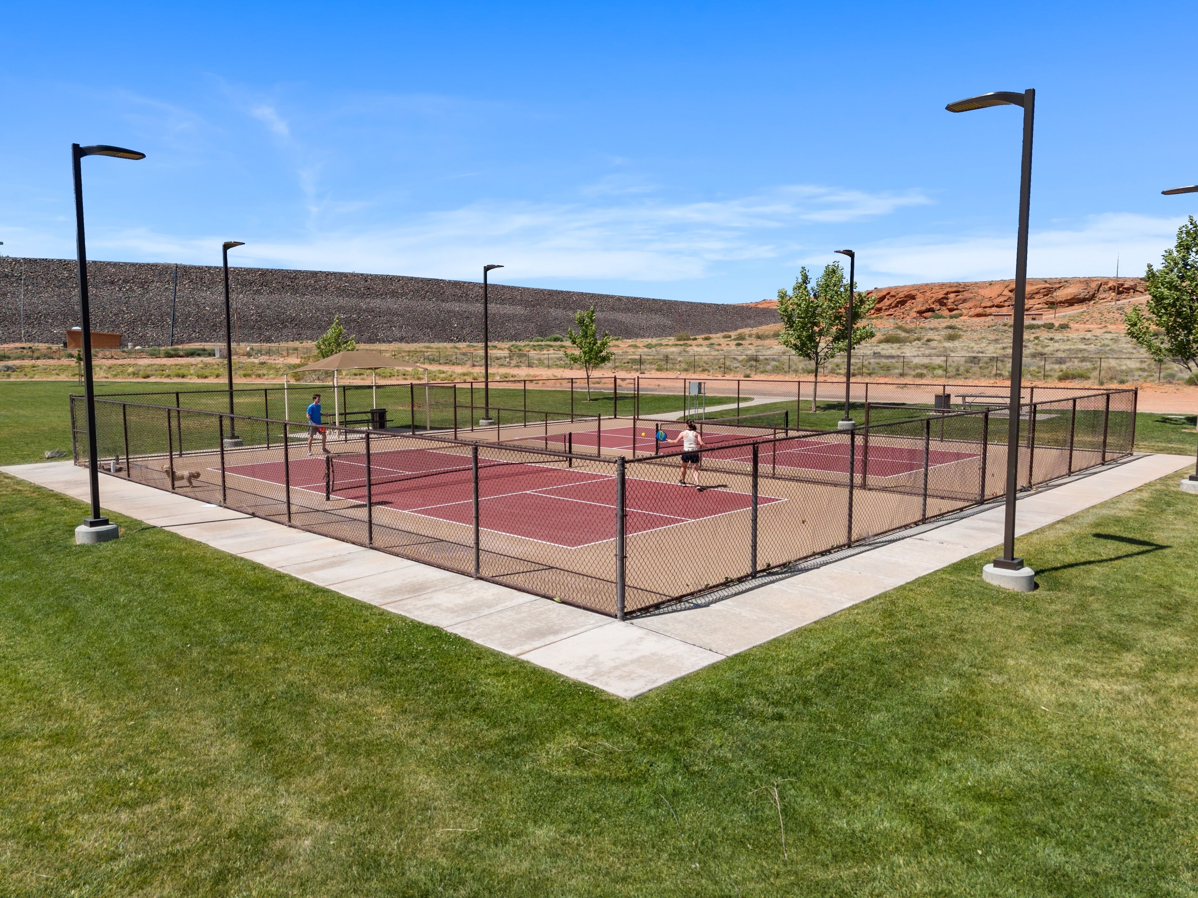 An outdoor tennis court with a chain-link fence, surrounded by grass and trees.