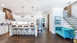 Une cuisine moderne avec des meubles blancs, un îlot central doté de tabourets de bar, des appareils en acier inoxydable et un escalier menant à un espace de vie agrémenté d’un canapé turquoise.
