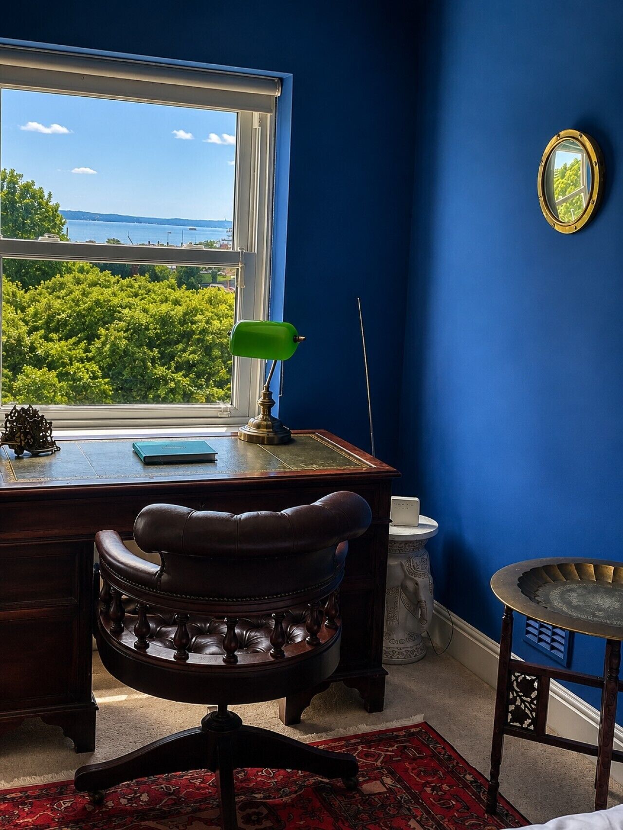 Una habitación con escritorio, silla y vista a follaje y agua a través de una ventana.