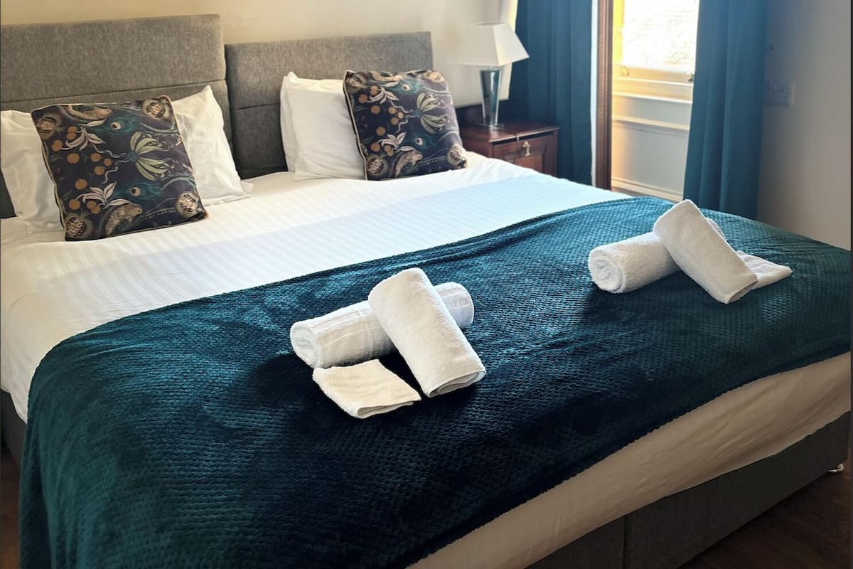 Ein ordentlich gemachtes Bett mit einer türkisfarbenen Decke und zwei aufgerollten Handtüchern, ein grauer Kopfteil und ein Nachttisch mit einer Lampe.