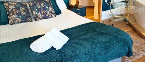 A neatly made bed with a teal blanket and pillows, a bedside table with a lamp, and a view of a bathroom with a bathtub and a towel rack.
