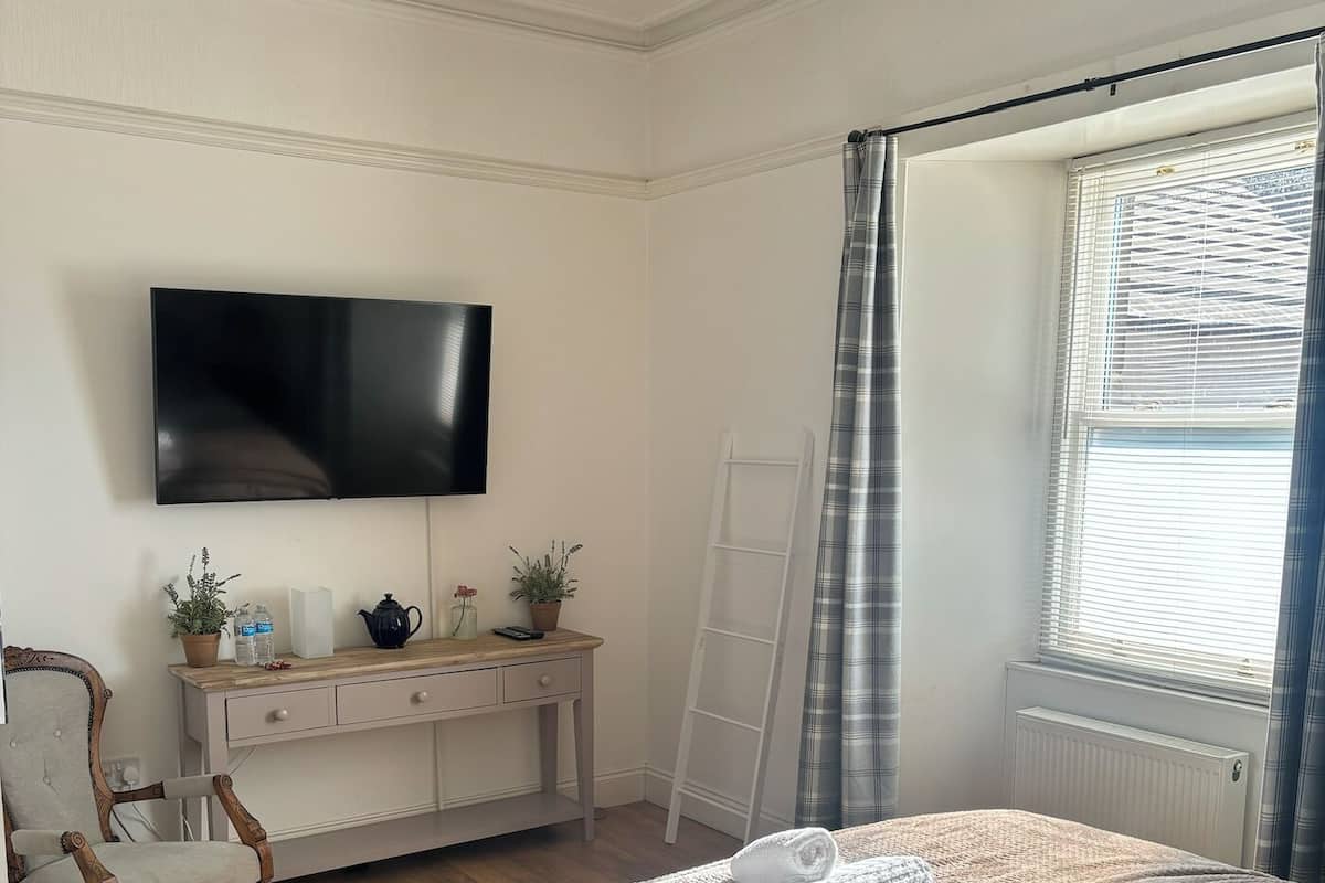 Ein Schlafzimmer mit Bett, einem an der Wand montierten Fernseher, einem Nachttisch mit Lampe, einem Sessel und einem Fenster mit Vorhängen.