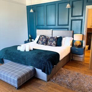 Quarto com cama grande, um banquinho, mesa de cabeceira e abajur.