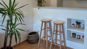 A kitchen with white cabinets, a hanging light fixture, two wooden stools, a potted plant, and a wall-mounted shelf with decorative items. - (New) Superb apartment in Golf (Saumane-de-Vaucluse)