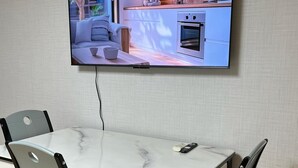 Una zona de menjador amb una taula i cadires, un televisor de pantalla plana penjat a la paret, i una cuina visible a través del televisor.