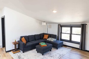 En moderne stue med en hjørnesofa, en lille skammel, et sofabord med en plante og et stort vindue med gardiner.