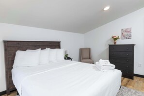Ein ordentlich bezogenes Bett mit weißen Leinen, ein dunkles Holz-Kopfteil, ein beiger Sessel, eine schwarze Kommode und ein kleiner Blumenstrauß auf der Kommode.