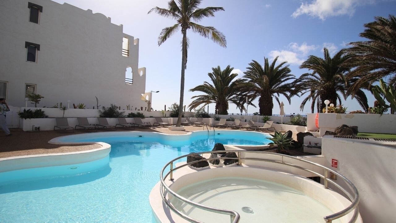 Un área de piscina con varias piscinas, palmeras y un edificio blanco con balcones.