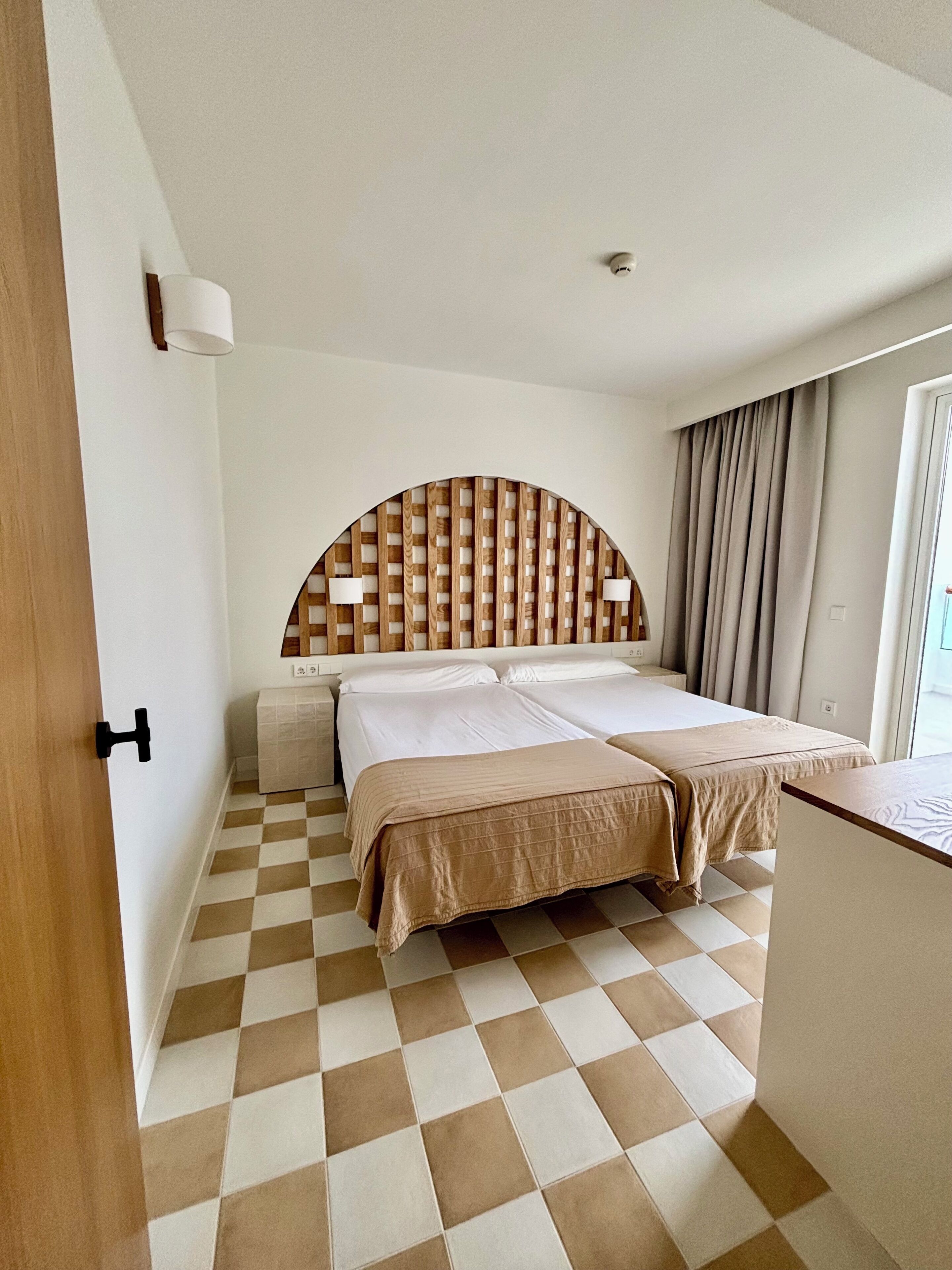 A hotel room with a checkered floor, a bed with a wooden headboard, a bedside table, and a window with curtains.
