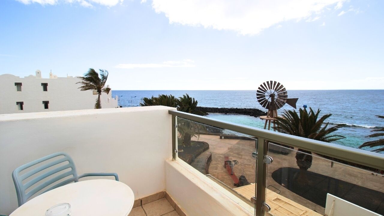 Un balcon avec une table et des chaises, donnant sur une plage où l’on aperçoit un moulin à vent et des palmiers.