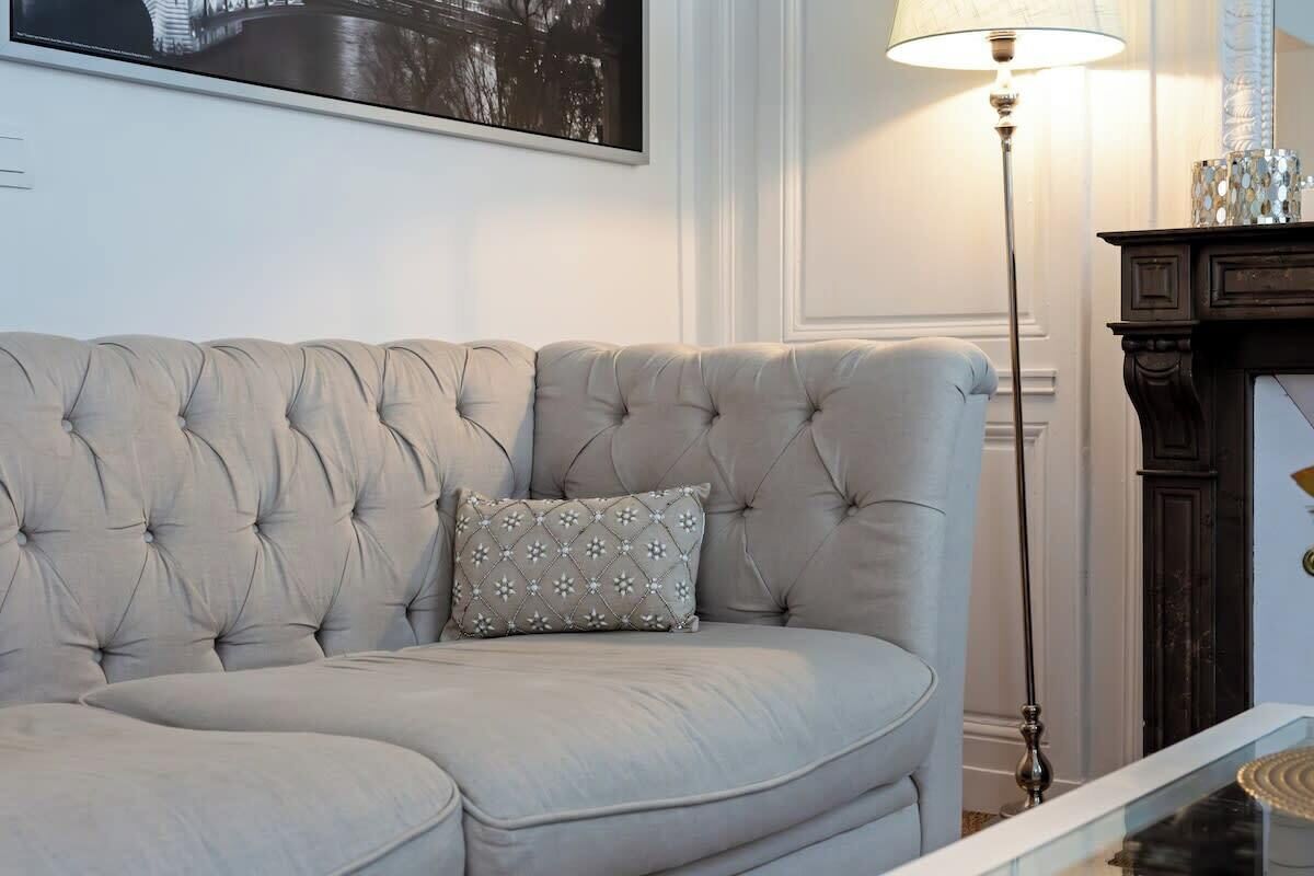 A light-colored, tufted sofa with a patterned pillow in a well-lit room with a floor lamp and a fireplace.