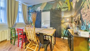 Un comedor con una mesa dispuesta para cuatro personas, rodeada de sillas coloridas, un suelo de madera y un mural en la pared con motivos de hojas.