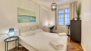 Un dormitorio con cama, mesita de noche, lámpara, escritorio, silla, radiador, ventana y una planta en maceta.