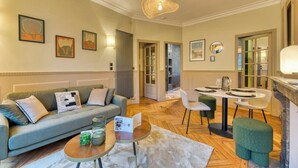 A modern living room with a sofa, coffee table, and dining area. There are framed pictures on the wall, a chandelier, and a fireplace. - Olivier : Élégance au Cœur de Rouen (Rouen)