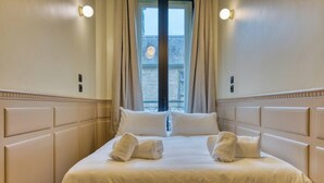 A neatly made bed with white linens and two towels, a window with curtains, and wall-mounted lights. - Olivier : Élégance au Cœur de Rouen (Rouen)
