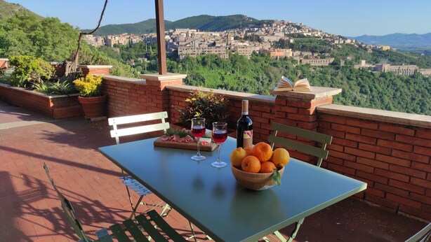 An outdoor patio with a table set for two, featuring a bowl of fruit, wine glasses, and a bottle of wine.
